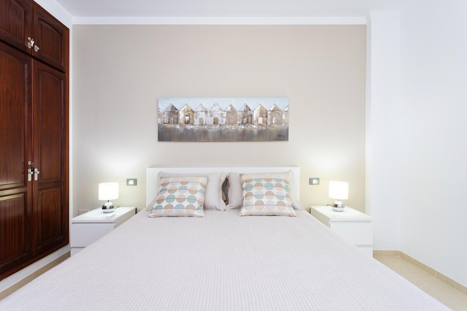 Un dormitorio con una cama grande, dos mesitas de noche, un armario de madera y un cuadro con una vista urbana en la pared.