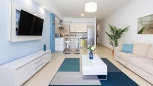Un salon moderne avec un téléviseur à écran plat, un canapé blanc, une table basse et une cuisine en arrière-plan.