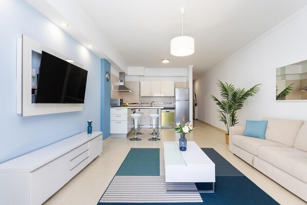 Un salon moderne avec un téléviseur à écran plat, un canapé blanc, une table basse et une cuisine en arrière-plan.