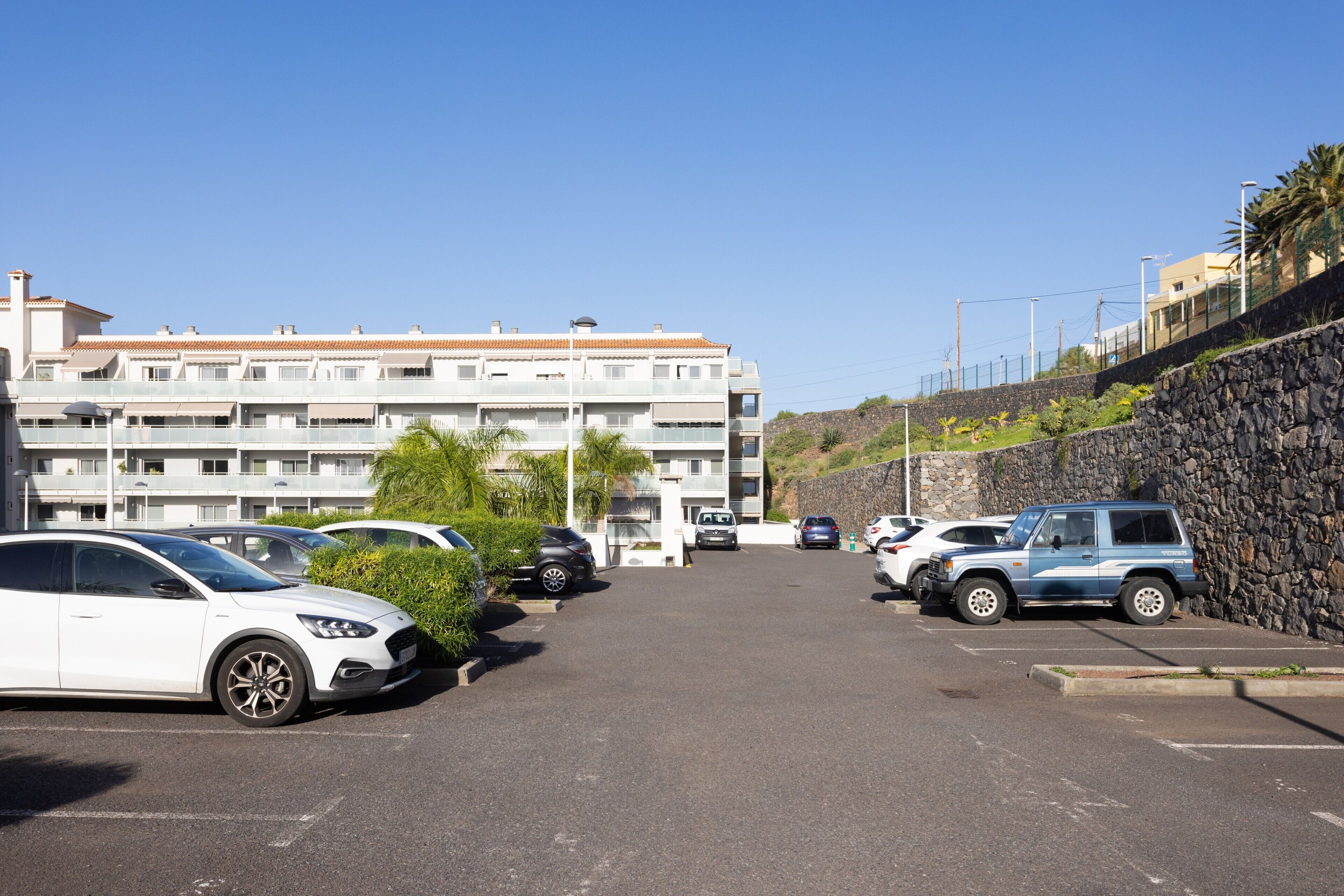 Un estacionamiento con varios autos, un edificio con múltiples balcones y un muro de contención de piedra.