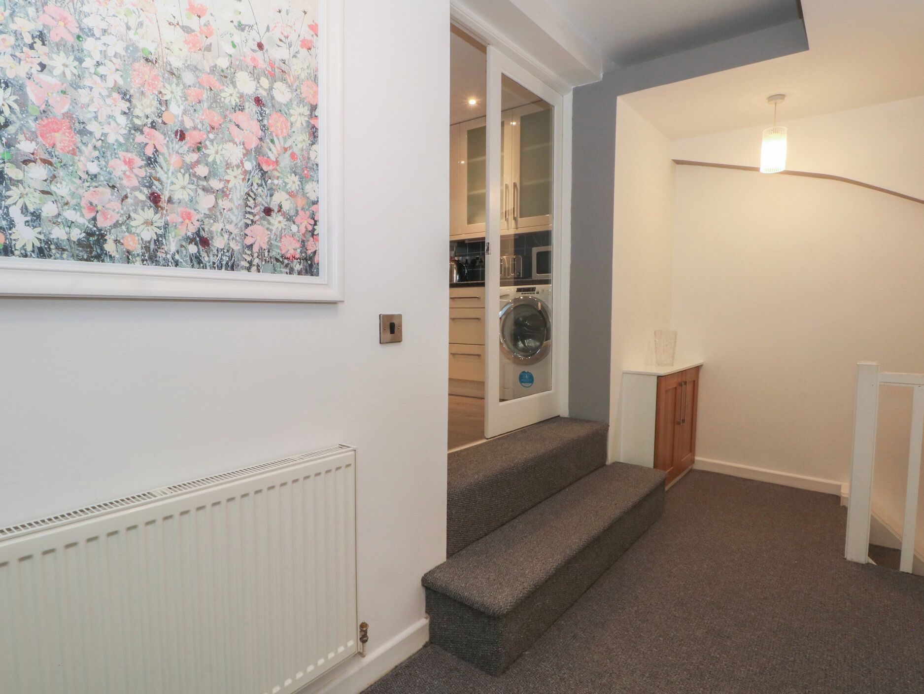 A staircase leading to a laundry area with a washing machine, a radiator, and a framed floral artwork on the wall.