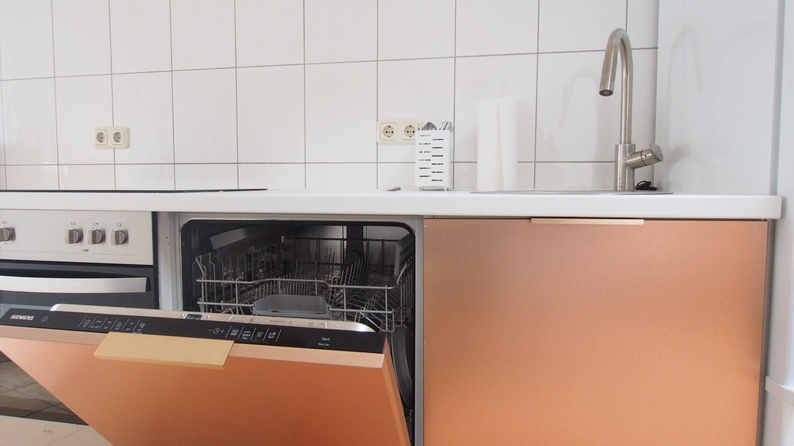 Cozinha com painel de azulejos brancos, pia de aço inoxidável e lava-louças.