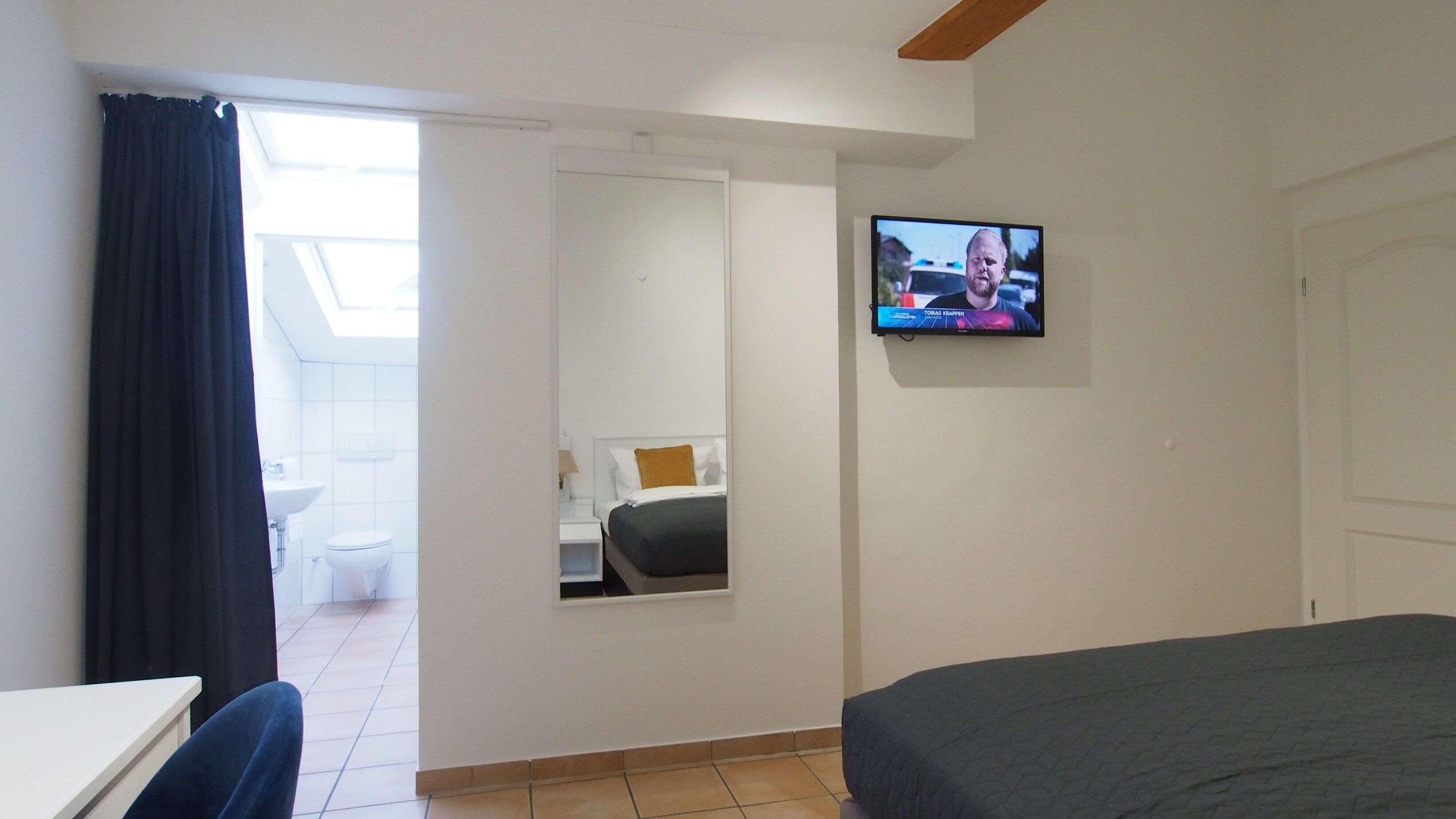 Habitación de hotel con cama, televisor, escritorio y baño visible a través de una puerta abierta.