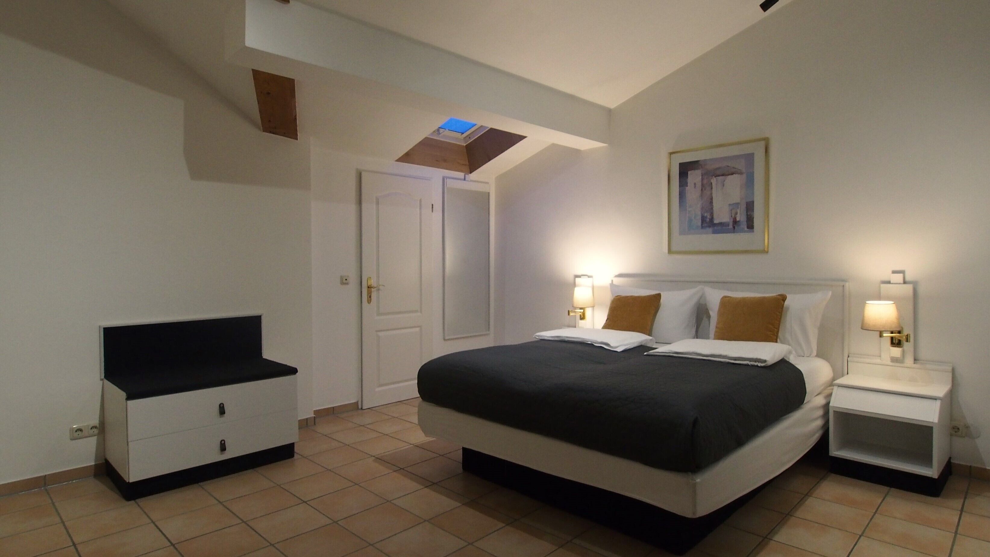 Un dormitorio con cama, mesitas de noche, armario, tragaluz y un cuadro en la pared.