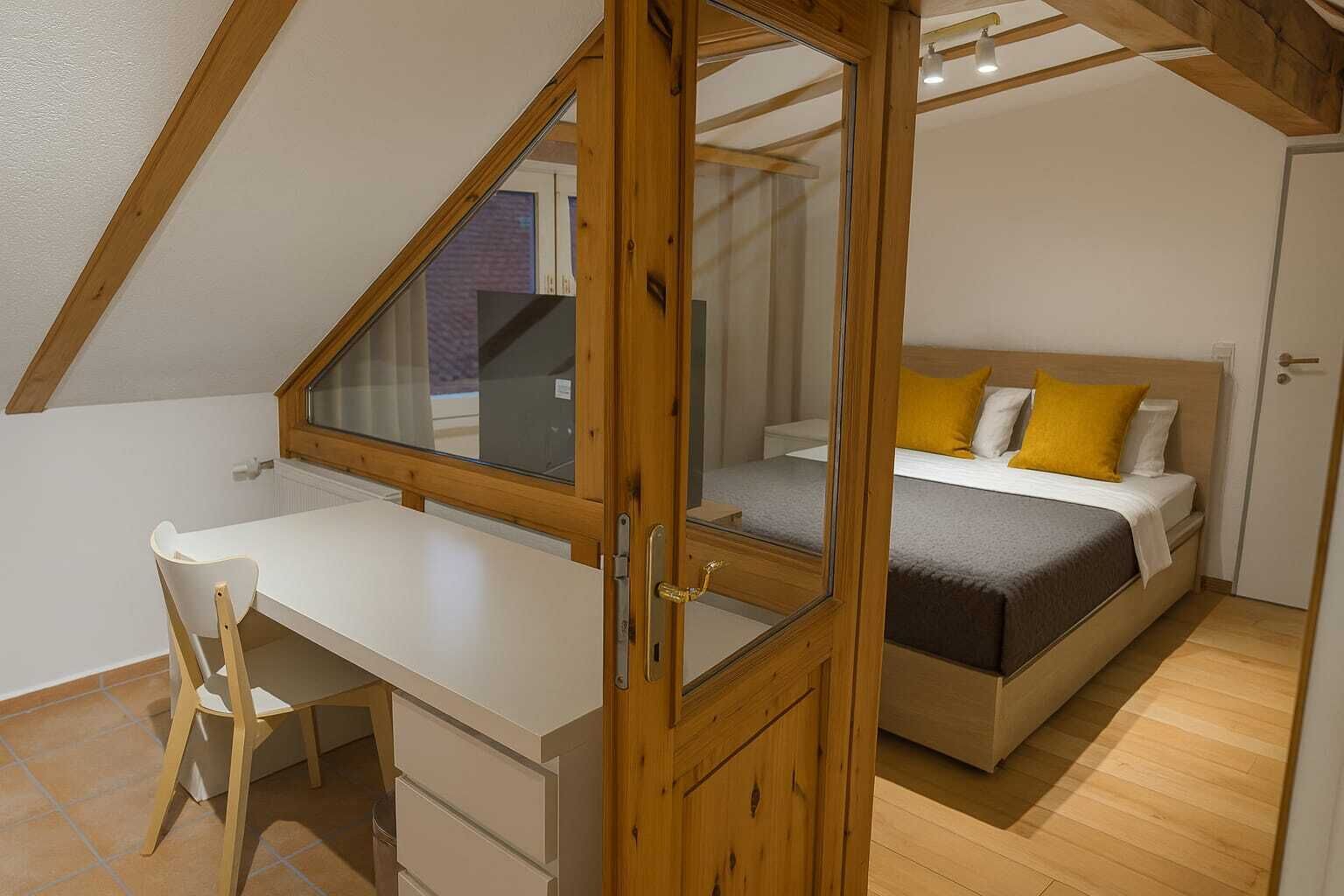 A bedroom with a sloped ceiling, a bed with yellow pillows, a desk with a chair, and a window.