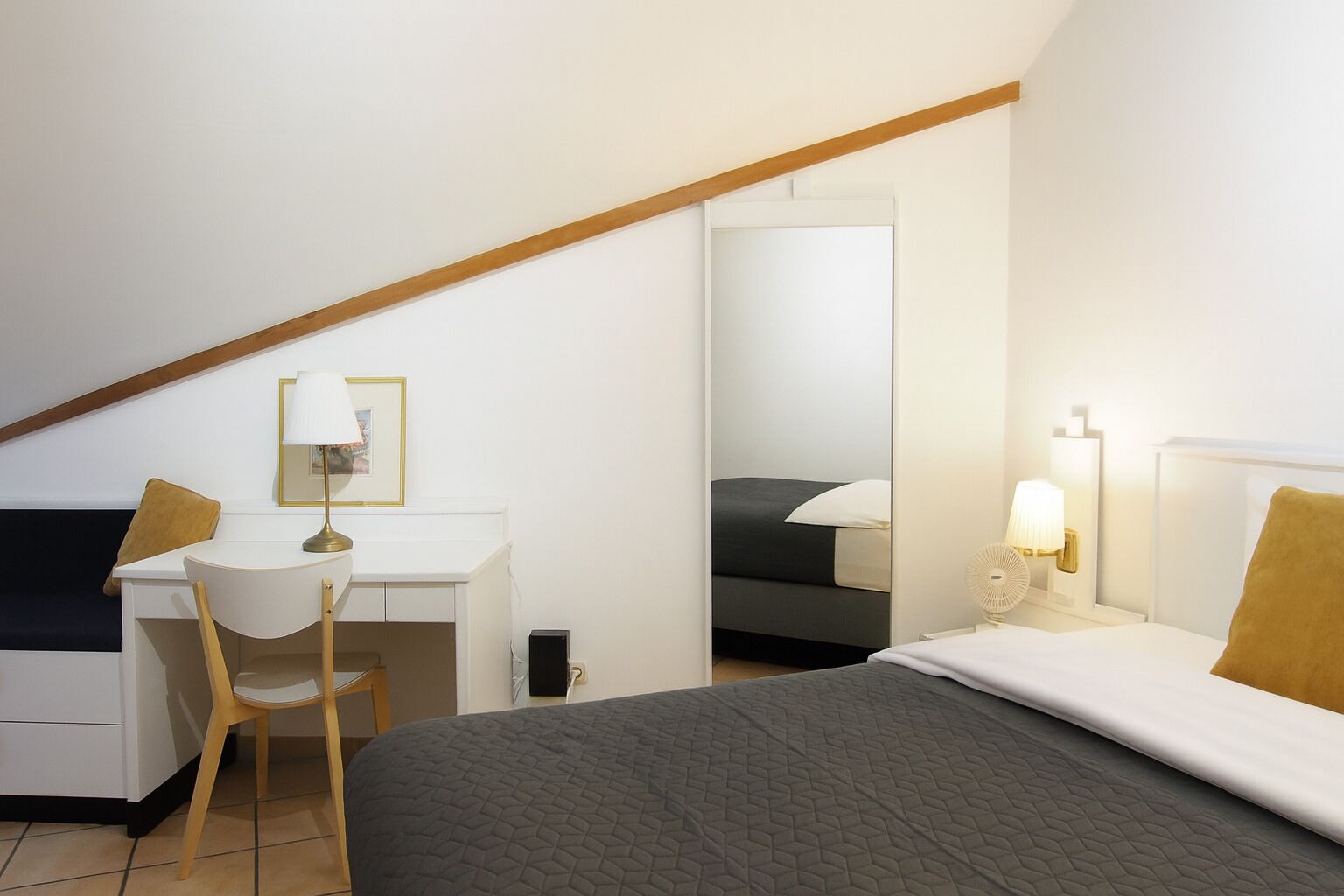A bedroom with a bed, a desk, a chair, and a nightstand with a lamp.