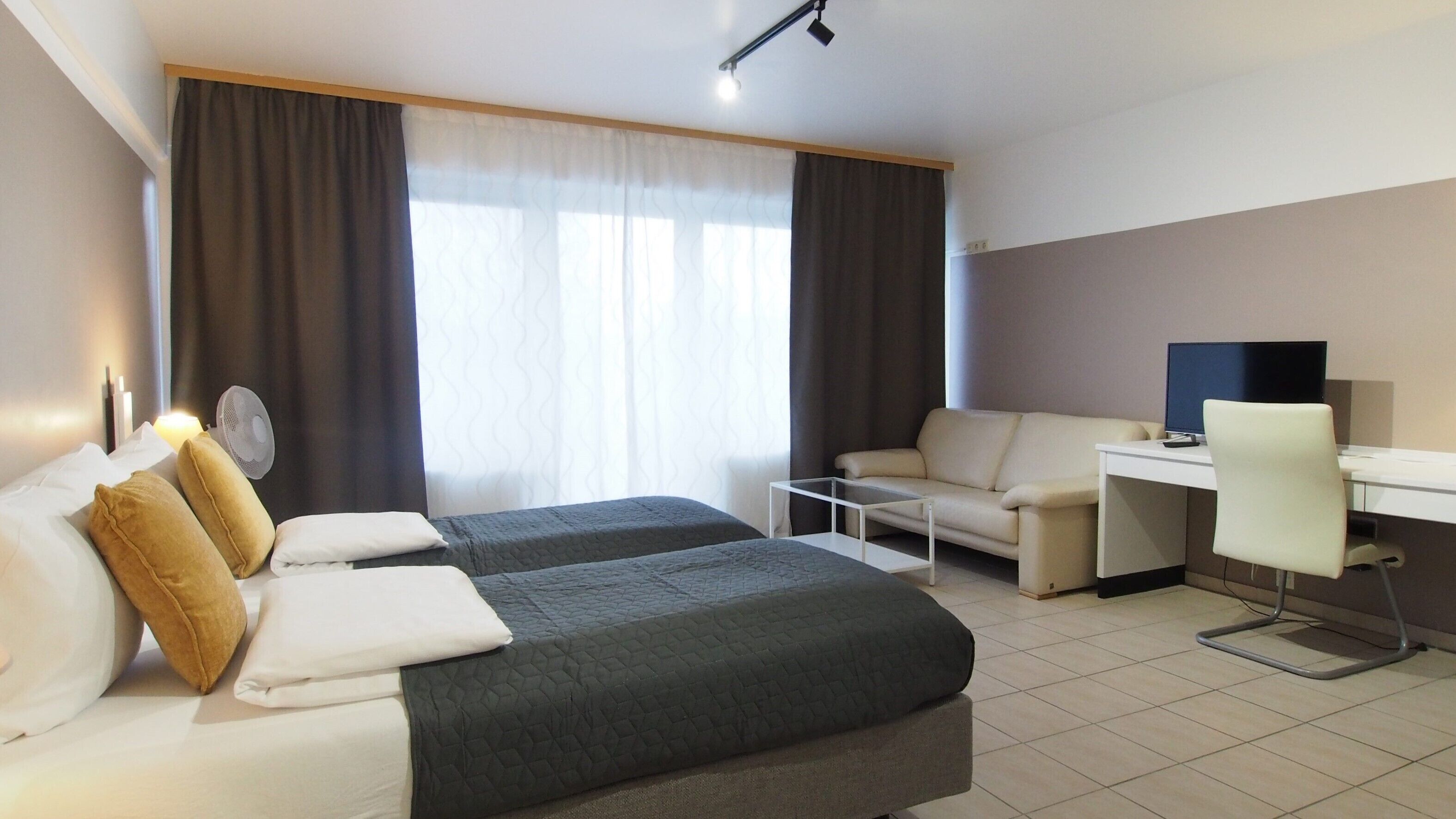 A hotel room with two beds, a desk, a chair, a TV, and a fan.