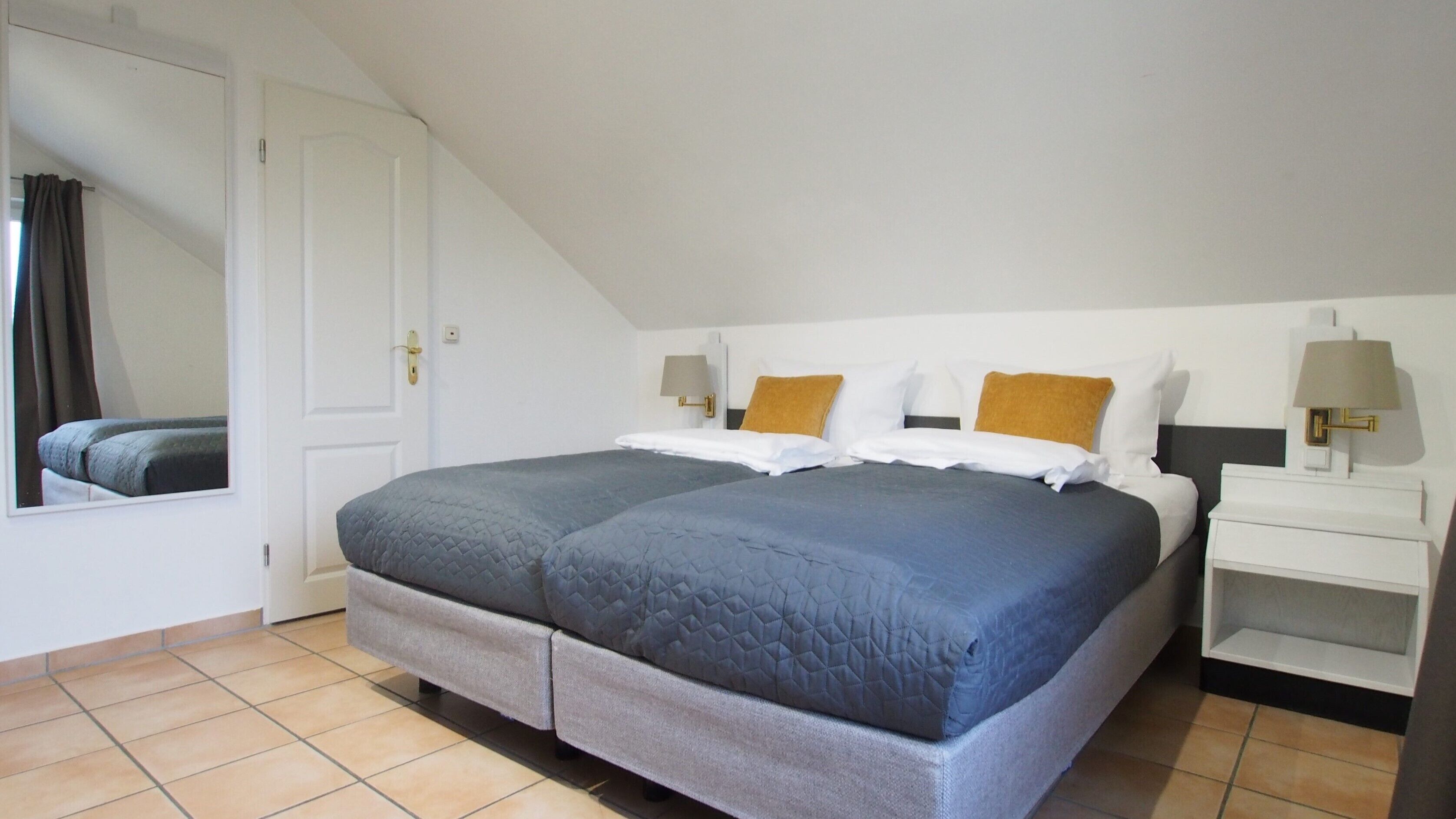 Een tweepersoonsbed met grijs en blauw beddengoed, twee nachtkastjes met lampen, een witte deur en een spiegel aan de muur.