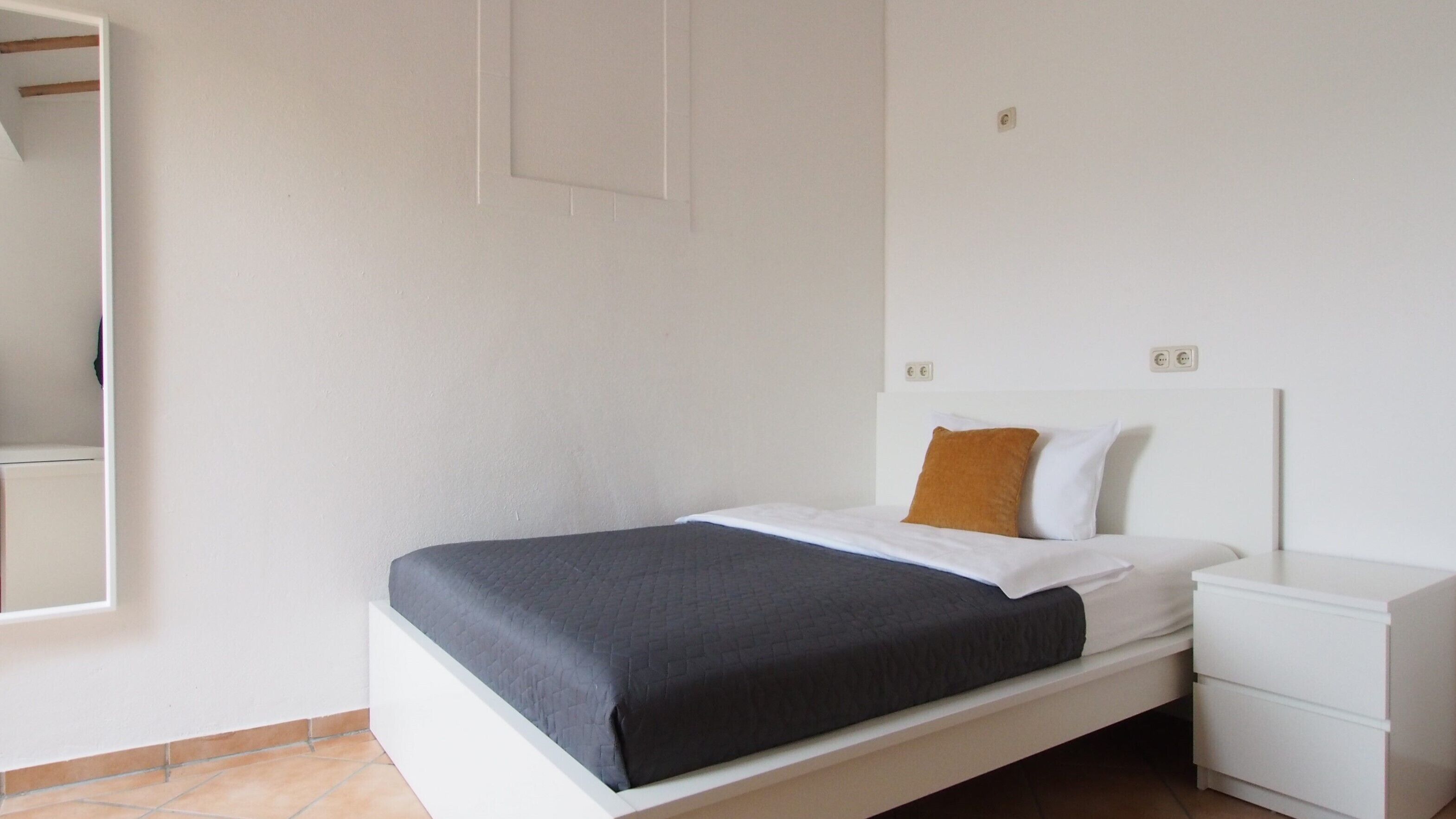 Un dormitorio minimalista con cama, mesita de noche blanca y espejo.