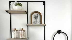 Een badkamer met een wandplank waarop een potplant, een lijst en twee glazen potten staan. Een handdoekhouder met een witte handdoek hangt rechts.