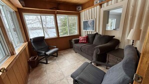 A cozy cabin interior with a wooden ceiling, a couch, two chairs, and a window overlooking a lake.