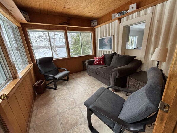 A cozy cabin interior with a wooden ceiling, a couch, two chairs, and a window overlooking a lake. - 4 season Family Friendly Beach Cottage in the Bay (Whitestone)