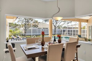 Een eethoek met uitzicht op een achtertuin en zwembad, met een tafel voor twee, stoelen en een hangende lamp.