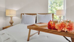 Una cama bien hecha con sábanas blancas, una mesita de noche de madera con un libro y flores, y dos lámparas de noche.