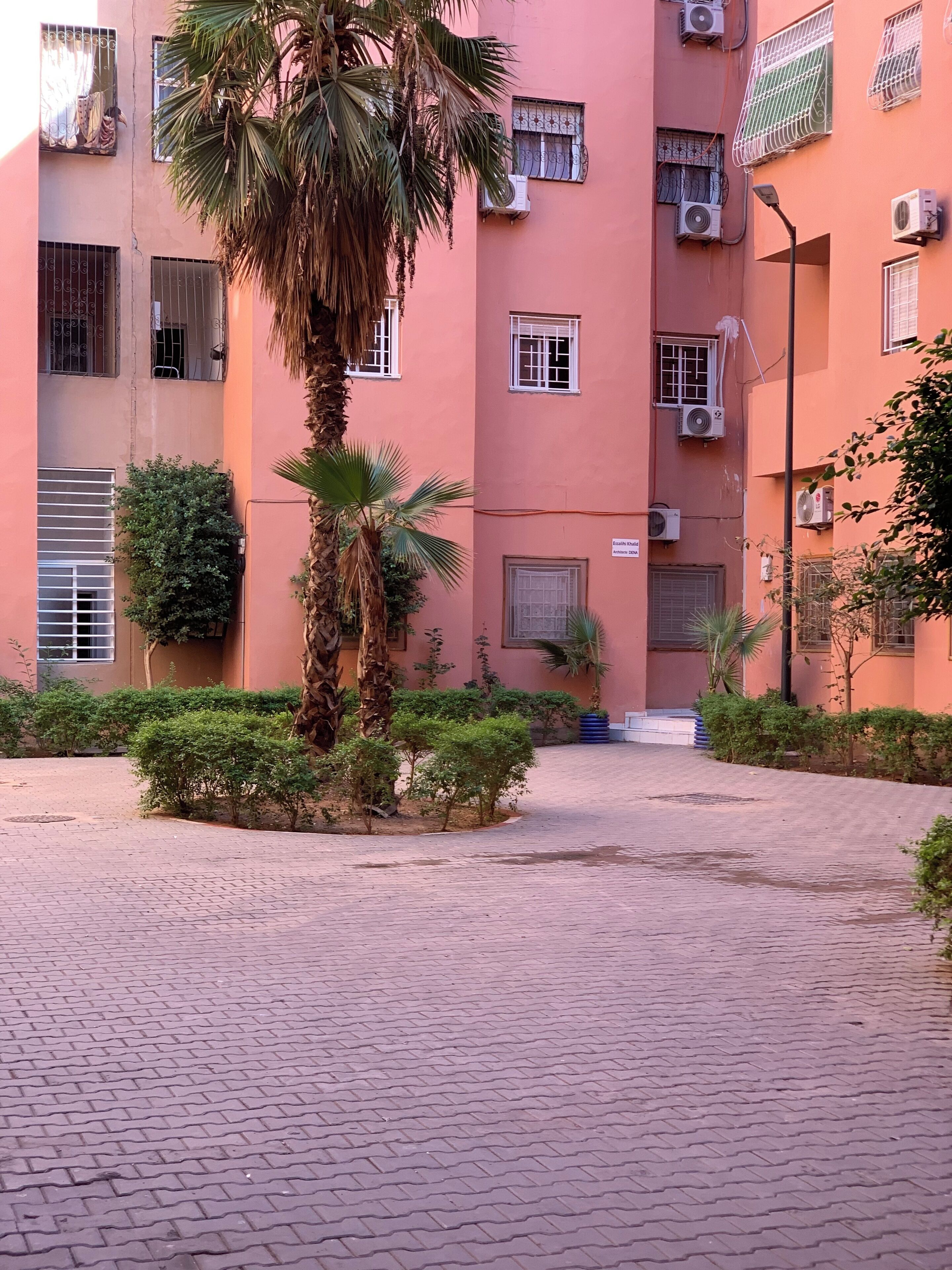 A residential building with multiple balconies, air conditioning units, and a paved courtyard with small trees and shrubs.