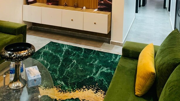 Ein modernes Wohnzimmer mit einem an einer Holzwand montierten Flachbildfernseher, einem grünen Sofa, einem Glastisch und einem Teppich mit Marmor-Muster.