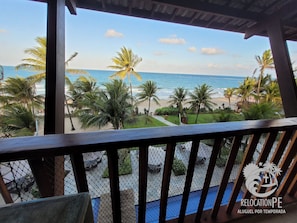 Uitzicht op het strand vanaf een overdekt balkon, met palmbomen en een heldere lucht.