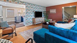 A living room with a blue sofa, a wooden cabinet, a television, and a kitchen area in the background. - Lake Escape-Pool Table-Kids Game Room-Wii-Foosball (Avon Lake)