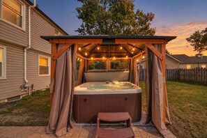 Un jacuzzi bajo una pérgola de madera con luces de cuerda, rodeado por un patio trasero con valla de madera y casas al fondo.