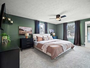 Un dormitorio con cama, mesitas de noche, ventilador de techo y ventana con cortinas.