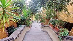 Un étroit sentier dallé, bordé de murs en briques et de plantes en pot, mène à un espace central.