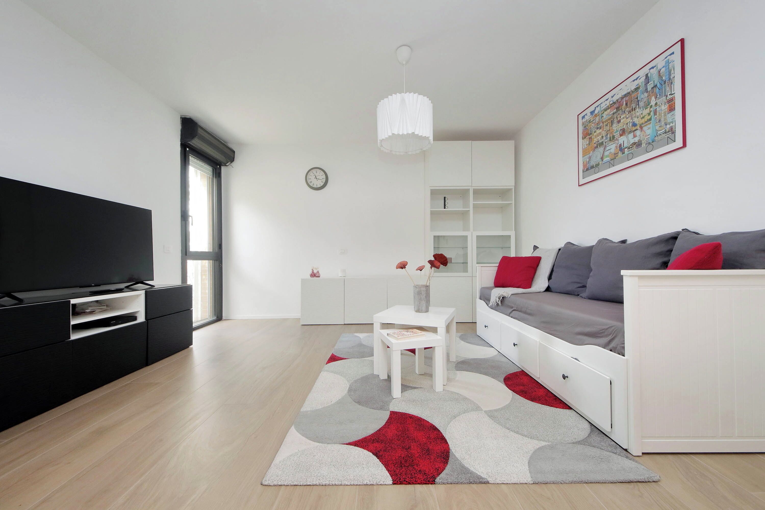 A modern living room with a flat-screen TV, a white sofa with red cushions, a white coffee table, and a large abstract rug.