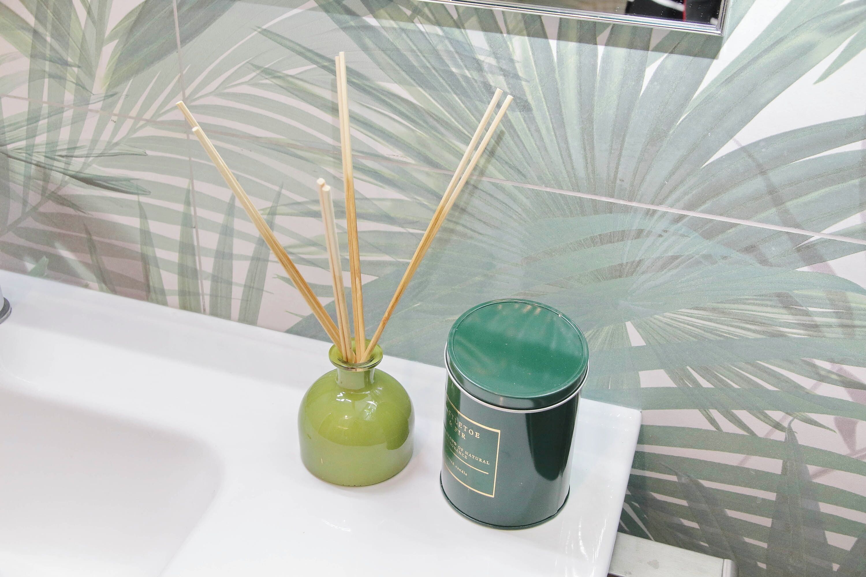 A bathroom with a glass partition, a green vase with reed sticks, and a green candle on the sink.