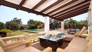 Zona d'estar exterior coberta amb mobiliari de vugada, una taula de cafè de vidre i vistes a una piscina i jardí.