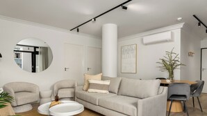 Ein modernes Wohnzimmer mit einem grauen Sofa, zwei Sesseln, einem runden Couchtisch und einem Esstisch mit Stühlen.