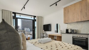 A modern hotel room with a large bed, a desk, a kitchenette, and a view of the city. - The York | Designer CBD Apartment (Cape Town)