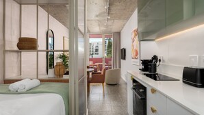 A modern hotel room with a bed, a small kitchenette, a seating area with a chair, and a view of the city through large windows. - Coastal Studio in the Heart of Sea Point | Pool (Cape Town)