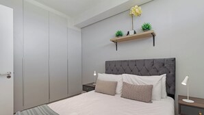 Une chambre à coucher bien aménagée, avec un grand lit, deux tables de chevet, une commode, une tête de lit rembourrée de couleur grise et une petite étagère avec des plantes.
