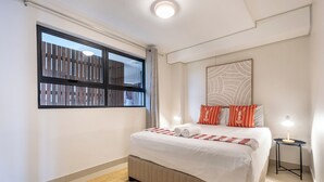 Ein Hotelzimmer mit einem großen Bett, einem Fenster mit Blick auf einen Holzzaun, einem kleinen Nachttisch mit Lampe und einem an der Wand hängenden abstrakten Kunstwerk.