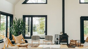 A spacious living room with a high ceiling, large windows, a fireplace, and a comfortable seating area. - Lakeside Calm at Dimensions | Cabin One | Breakfast Included (Algonquin Highlands)