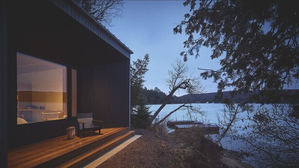 A modern cabin with a large window overlooking a lake, a wooden deck, and a small outdoor seating area. - Lakeside Calm at Dimensions | Cabin One | Breakfast Included (Algonquin Highlands)