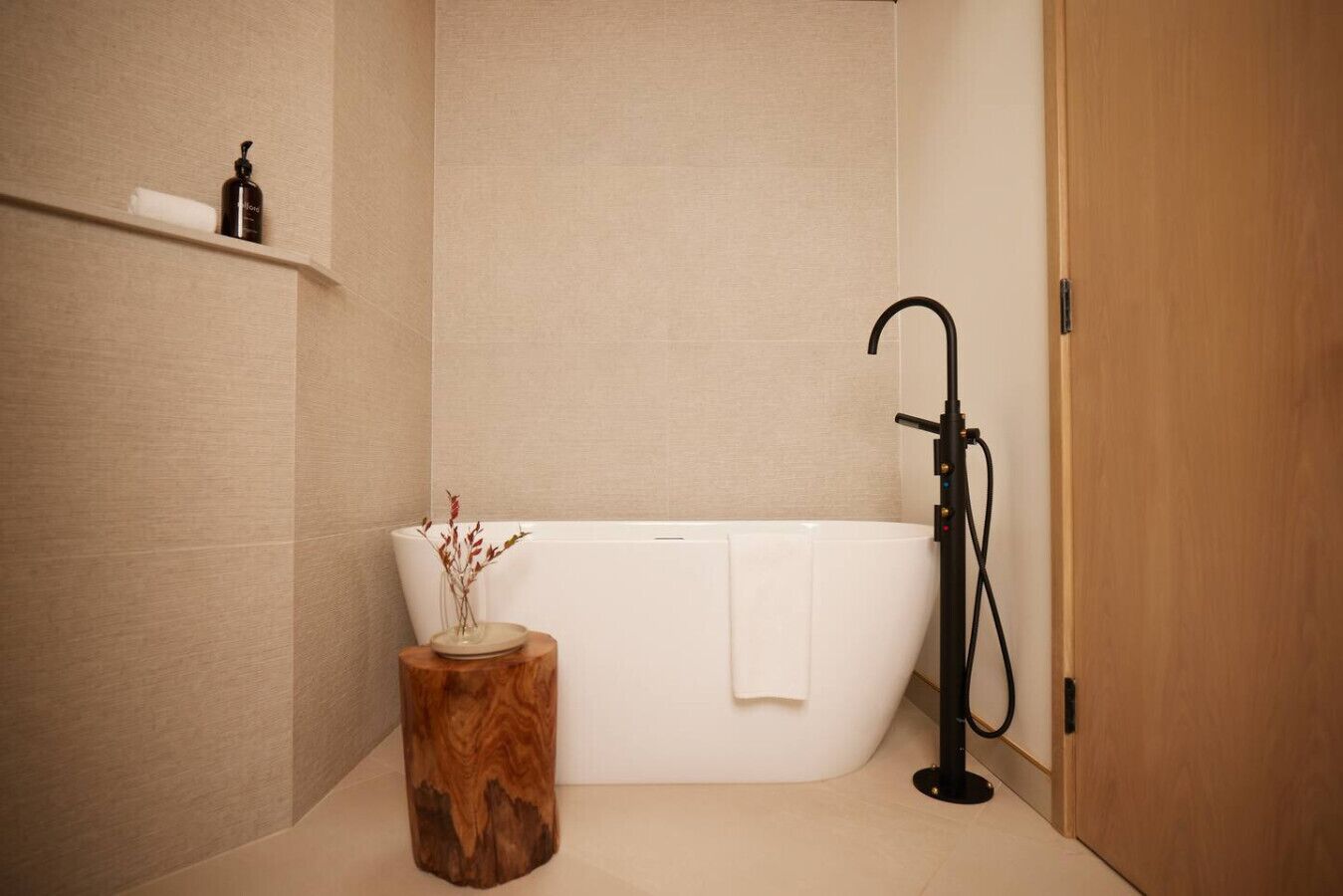 Een moderne badkamer met een vrijstaand bad, een houten bijzettafeltje met een vaas, en een plank met toiletartikelen.