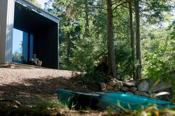 Un edificio moderno con un gran ventanal, rodeado de zona boscosa con una terraza de madera y un kayak.