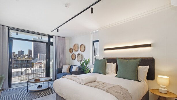 A modern hotel room with a large bed, a small round table, a balcony with a city view, and a wall-mounted lamp. - Cosy Studio Apartment | The York (Cape Town)