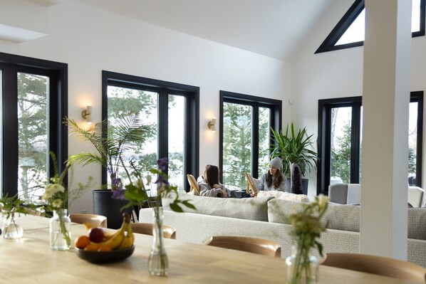 A modern living room with large windows, a dining table with fruit, and a sofa.