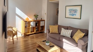 Ein Wohnzimmer mit braunem Sofa, einem Holztisch, einem Korbstuhl, einer Holzkonsole mit Vase und Pflanze sowie einem gerahmten Bild an der Wand.