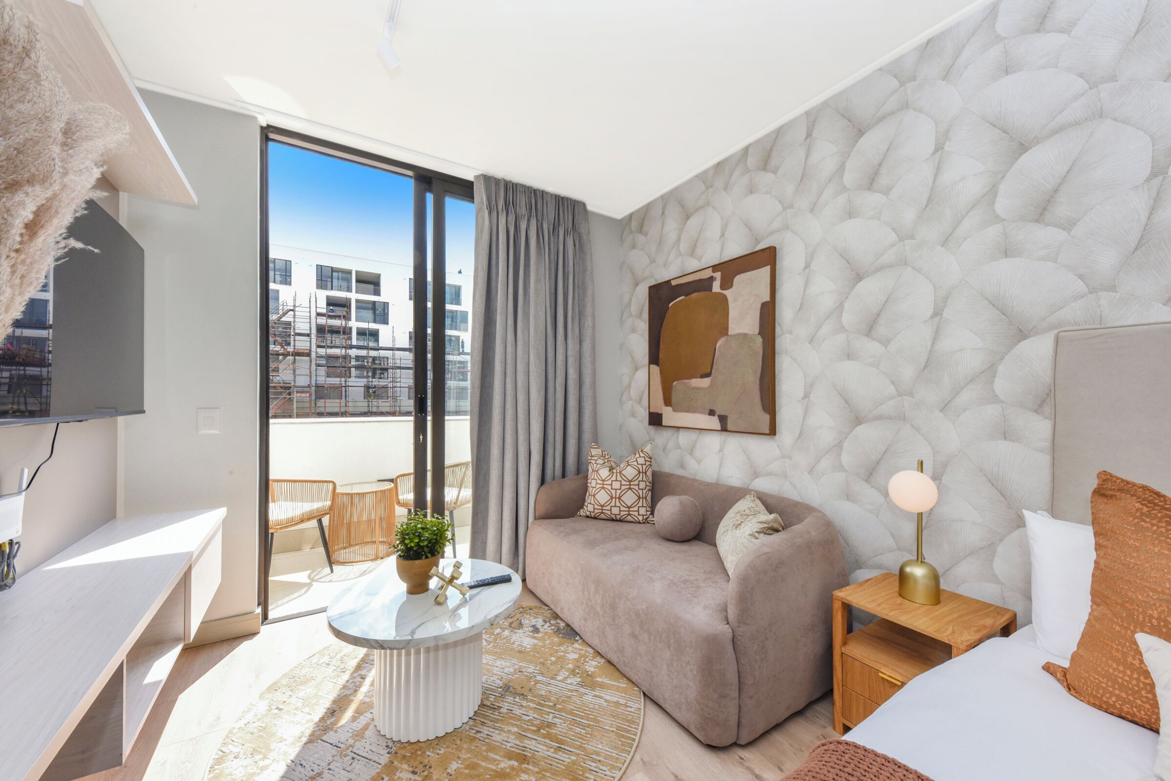 Quarto de hotel moderno com sofá, mesa de cabeceira, abajur e vista da cidade através de grandes janelas.