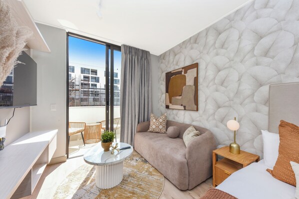 Quarto de hotel moderno com sofá, mesa de cabeceira, abajur e vista da cidade através de grandes janelas.