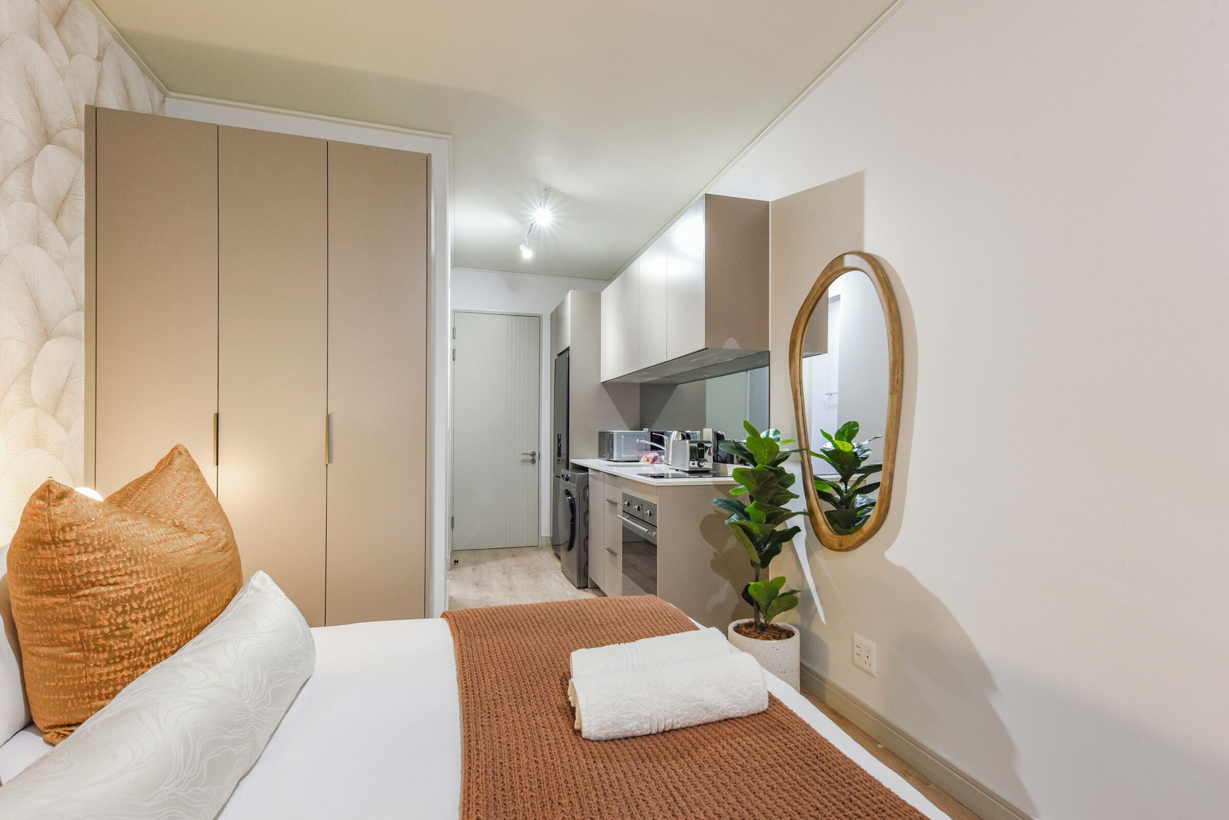 A hotel room with a bed, a large mirror, a potted plant, and a kitchenette in the background.