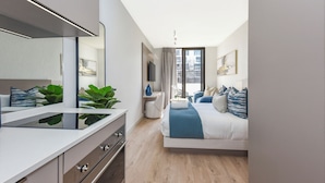 A modern hotel room with a large bed, a desk with a flat-screen TV, a mirror, and a view of the city through a large window. - The Velvet Touch | One Rosebank Studio (Johannesburg)