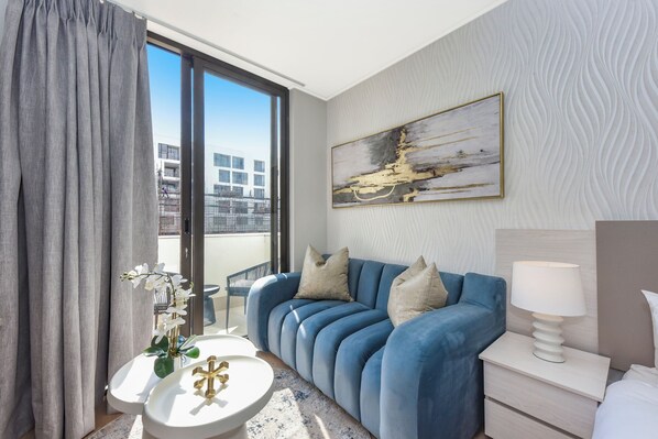 Une chambre d’hôtel moderne avec un canapé bleu, une table de chevet blanche, une petite table et une grande fenêtre donnant sur un immeuble.
