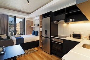 Habitación de hotel compacta con kitchenette, cama con almohadas azules, un pequeño comedor y vista a la ciudad.