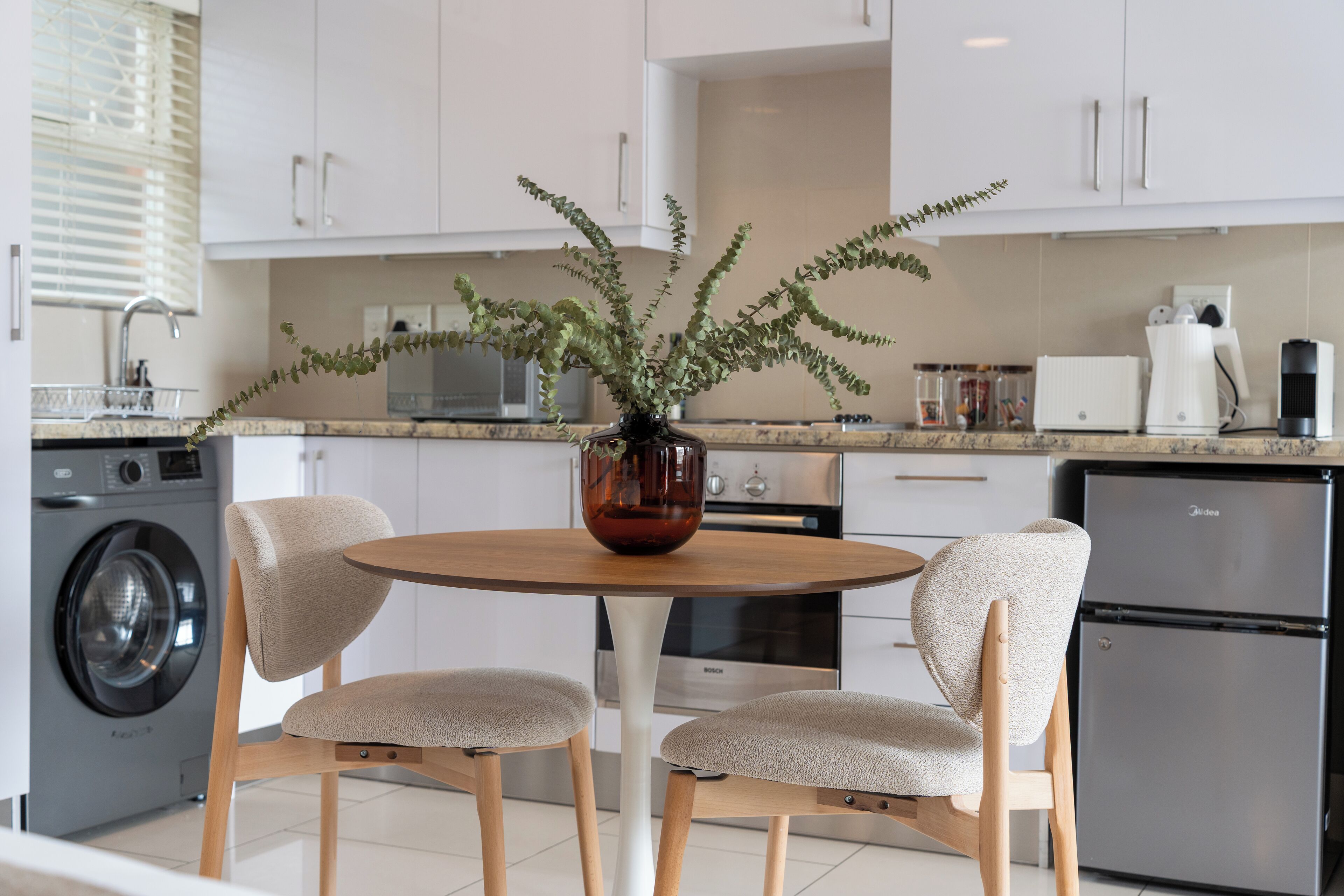 Une cuisine moderne avec des meubles blancs, une table ronde en bois, deux chaises beiges, un lave-linge, un réfrigérateur et une machine à café.