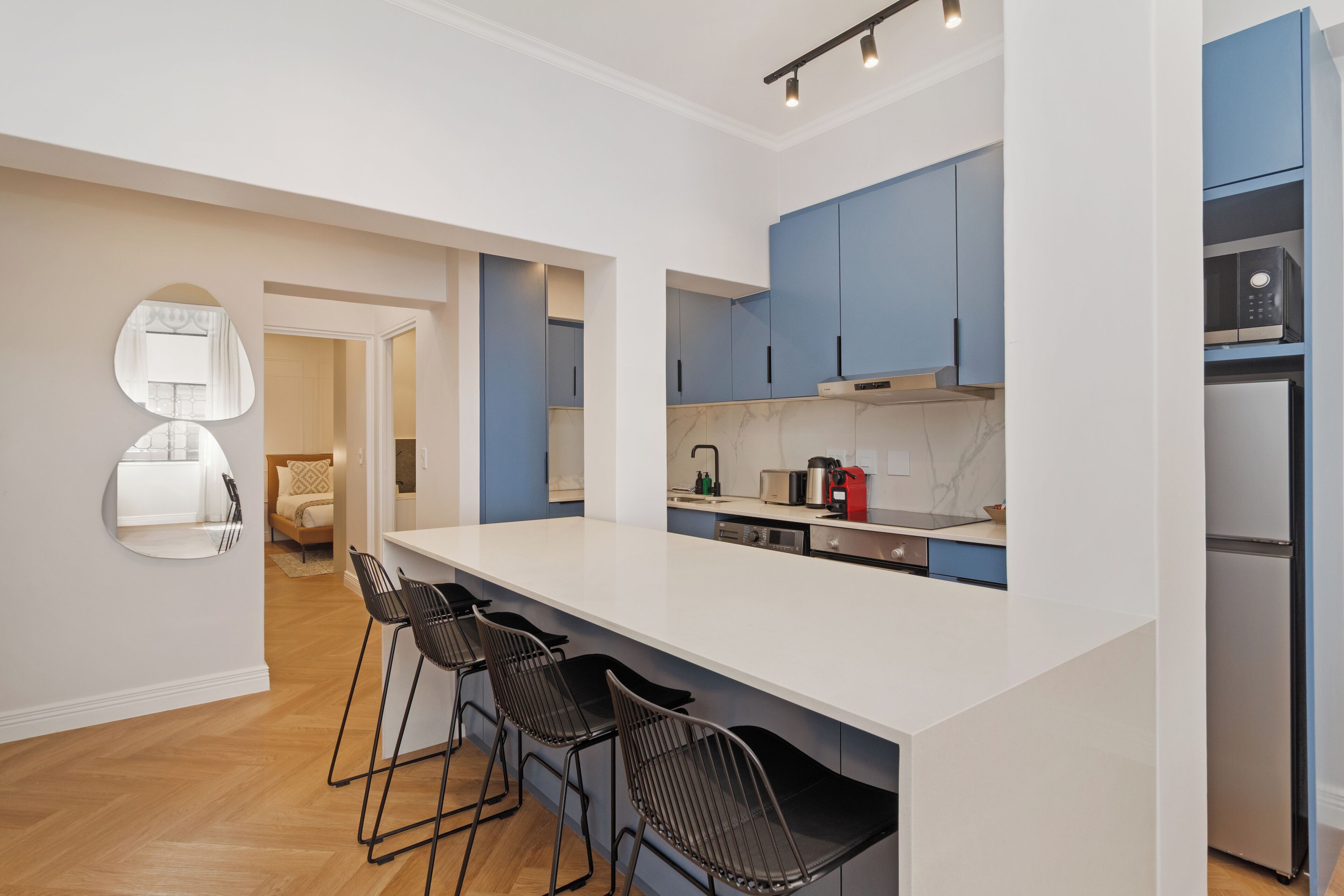Eine moderne Küche mit weißer Arbeitsplatte, blauen Schränken und einer eingebauten Mikrowelle. Drei schwarze Barhocker und ein großer Spiegel sind an der Wand angebracht.