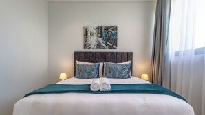 Cama bem arrumada com manta azul-turquesa, duas almofadas com padrão azul e dourado, e cabeceira com pintura preta e branca de uma bicicleta.