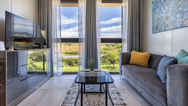 Uma sala de estar moderna com mesa de centro de vidro, sofá cinza com almofadas, televisão de tela plana e uma grande janela com cortinas.